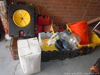 Photo showing boxed Subaru Outback car cover, six red gas cans, Petmate pet feeding systems, orange bucket, disposable garage rags, and squeegee stored on crates and a bin.
