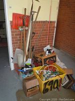 Photo of assorted yard tools including rakes, hoes, hedge trimmers, boxes, bins with extension cords and nails, and various poles and rods leaning against a wall.