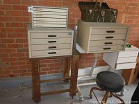 Photo showing two medium to large metal craft cabinets with drawers on wooden stands, a black round cushioned stool, and a metal toolbox with vintage spirit level on top.