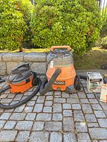 Two RIDGID shop vacuums with hoses, boxed filter and bags on cobblestone surface