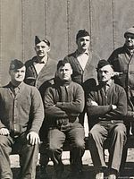 front of photograph showing 5 seated and 6 standing internees, wooden wall backdrop, placard 62 visible