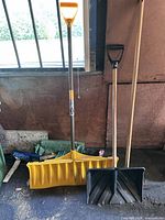 All three snow shovels standing: yellow wide pusher, black square shovel, wood-handled pusher