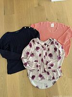 Three tops laid out: navy sweater, coral sweater, floral blouse