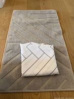 Folded white shower curtain with grey pattern placed on top of light grey textured bath mat
