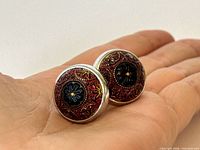 Front view of cufflink faces displaying red enamel, floral motif, size reference with hand