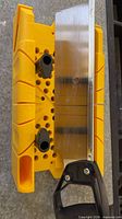 Top view of yellow miter box with saw inserted, showing black pegs and blade