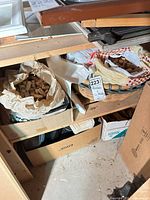 Shelf unit holding two plastic bags filled with corks, one bag inside a wicker basket; additional wicker basket, shelves, wood legs and cardboard boxes visible