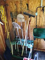 Overview of assorted long-handled garden tools in rack with spreader hopper and fencing roll