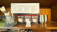 Shelf view showing three Make Market mug sets, Cricut 6-pack mug box, two stainless tumblers, mesh utensil cup