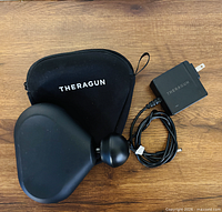 Theragun Mini massager with case and charger laid out on wooden surface