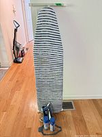 Ironing board standing upright with striped cover, Rowenta iron, two spray cans and silicone rest at base