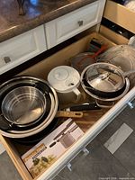 Drawer view showing stacked pans, bowls, boxed potato ricer, canister, grater and cake pans