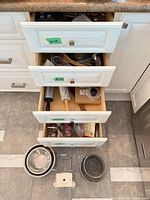 Three open kitchen drawers showing wide view of knives, rolling pins, whisks, tongs, mixing bowls and slicer base