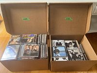 Two open cardboard boxes filled with numerous CD jewel cases; identifiable covers Lonestar, Il Divo, Good Old Country