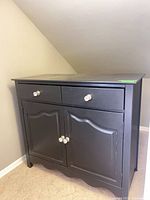 Full view of black storage cabinet showing two drawers, two doors, clear knobs, scalloped base