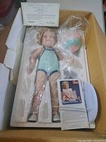 Doll in original styrofoam with plastic wrap, certificate, booklet visible