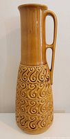front view of tall gold ceramic vase with handle and scroll relief