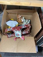 Open box showing assorted contents including flowers, telescope, figurines, Coca-Cola plane