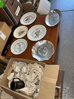 Overview of six plates on table and two mugs in packing box