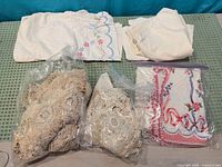 All linens laid out: embroidered tablecloth, plain cloth, two bags crochet doilies, bag embroidered napkins