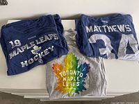 Two navy and one grey Toronto Maple Leafs T-shirts folded, showing Lupul front print, Matthews back print, grey rainbow logo