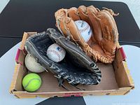 Overview of both gloves inside cardboard tray with two baseballs, one softball, one tennis ball