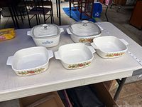 Five square CorningWare dishes, two with lids, displayed on table with measuring tape