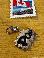 Front view of sterling silver POP charm with lobster clasp beside postage stamp for scale