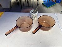 Two amber glass saucepans and marble mortar with pestle on table, measuring tape for scale
