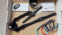 All items laid in box: Polar HRM wrist unit, chest strap and box, black Swatch watch with booklet, six multicolour Fitbit bands