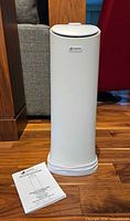 Front view of white tower humidifier with logo badge and manual on floor