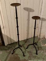 Pair of wrought iron floor candle holders standing against wall, showing overall form and height difference
