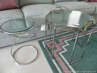 Pair of glass top accent tables on a patterned rug in front of a couch: one octagonal chrome table with brass handles and one round chrome spiral base table.