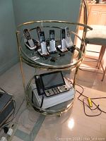 Front and side views of the round two tier glass table with gold and nickel toned metal frame showing multiple cordless phones and a landline phone on different shelves.