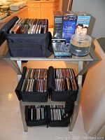 Overall view of metal stand with three tempered glass shelves holding four black fabric holders filled with CDs and sealed DVD box sets on top shelf.