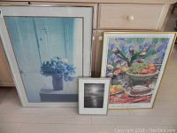 Three framed art pieces including two still life prints and one black and white seascape photograph arranged side by side on the floor in front of a dresser.