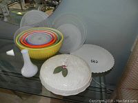 Photo showing a set of multicolored ceramic nesting mixing bowls, a white Lazy Susan, a white apple pie dish lid with 3D apple and leaves, and a small white vase on a glass table.