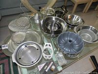 View of entire assortment of kitchenware including mixing bowls, colanders, trays, teapot, and baking accessories on a glass table.