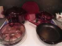 Full set showing two purple Vision glass pots with lids, black frying pan with red handle, and round ribbed glass dish with lid.