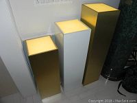 Front view of three lighted pedestals showing illuminated tops; two brass finish and one white, square bases.