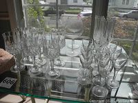 Overall view of 20 glassware pieces arranged on glass shelf near window including sets of wine glasses and goblets of different styles and sizes.