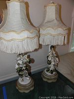 Pair of ornate table lamps with fabric fringe shades and ceramic figurine bases depicting a man and woman in traditional Asian dress, positioned indoors near a couch.