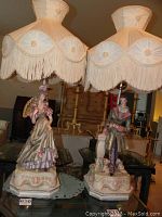 Both Victorian style table lamps with porcelain figurines of a man and woman with fabric fringe lampshades shown side by side on a table.