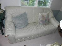 Front view of off-white leather couch with two throw pillows on a light-colored hardwood floor in front of windows.
