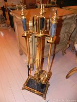 Full view of brass fireplace tool set with marble handles and matching marble base standing in front of cream colored cabinet.