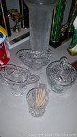 Image of assorted crystal dishes including a tall textured vase, a covered candy dish, a heart-shaped bowl, and a toothpick holder filled with toothpicks, showing cut glass patterns and clear transparency.