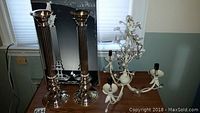 Two tall Godinger Silver Art silver-tone candlesticks standing next to a white floral chandelier on a wooden surface. The candlesticks appear unused in original box.