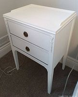 Front angled view of white two-drawer nightstand showing brass pulls and applique details