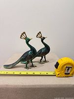 Front angled view of both brass enamel peacocks with tape measure for scale