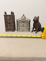 Front view of three cast iron coin banks: two buildings and seated dog with tape for scale